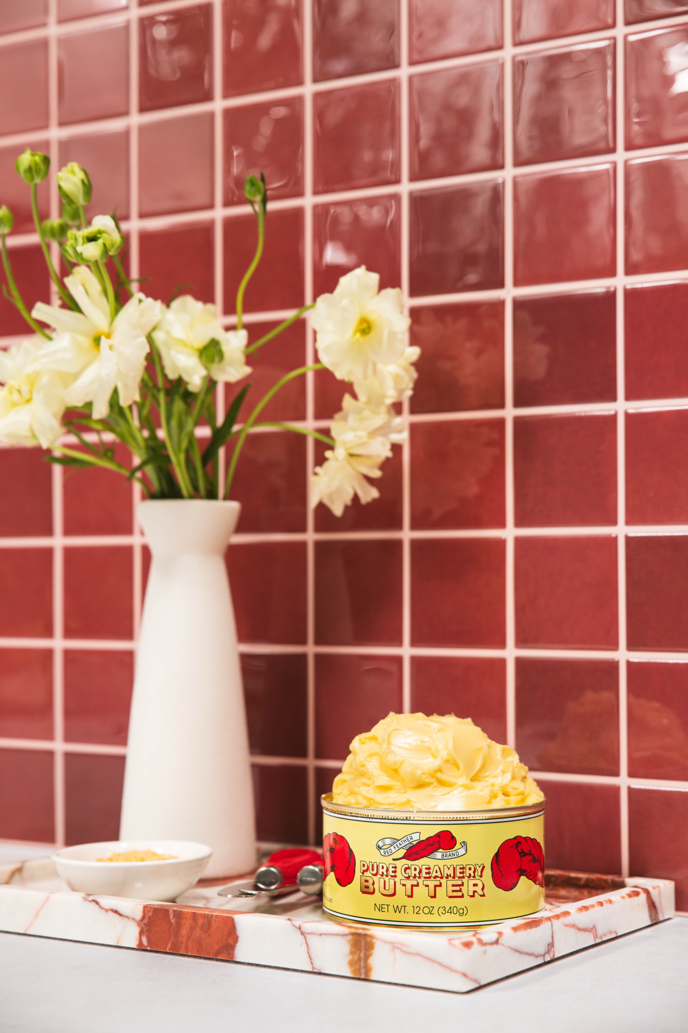 A tin of Red Feather butter on a marble slab with fresh flowers against a red tiled wall