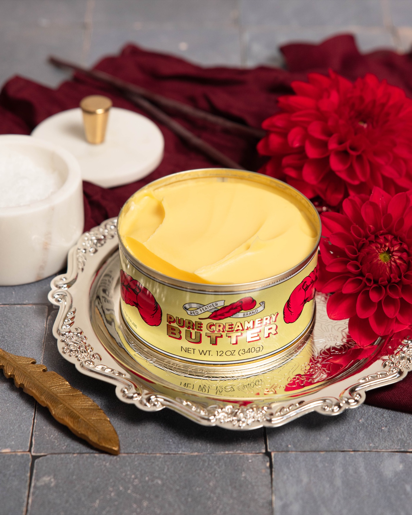 An opened tin of Red Feather butter on a silver tray with a brass knife and a red dahlia