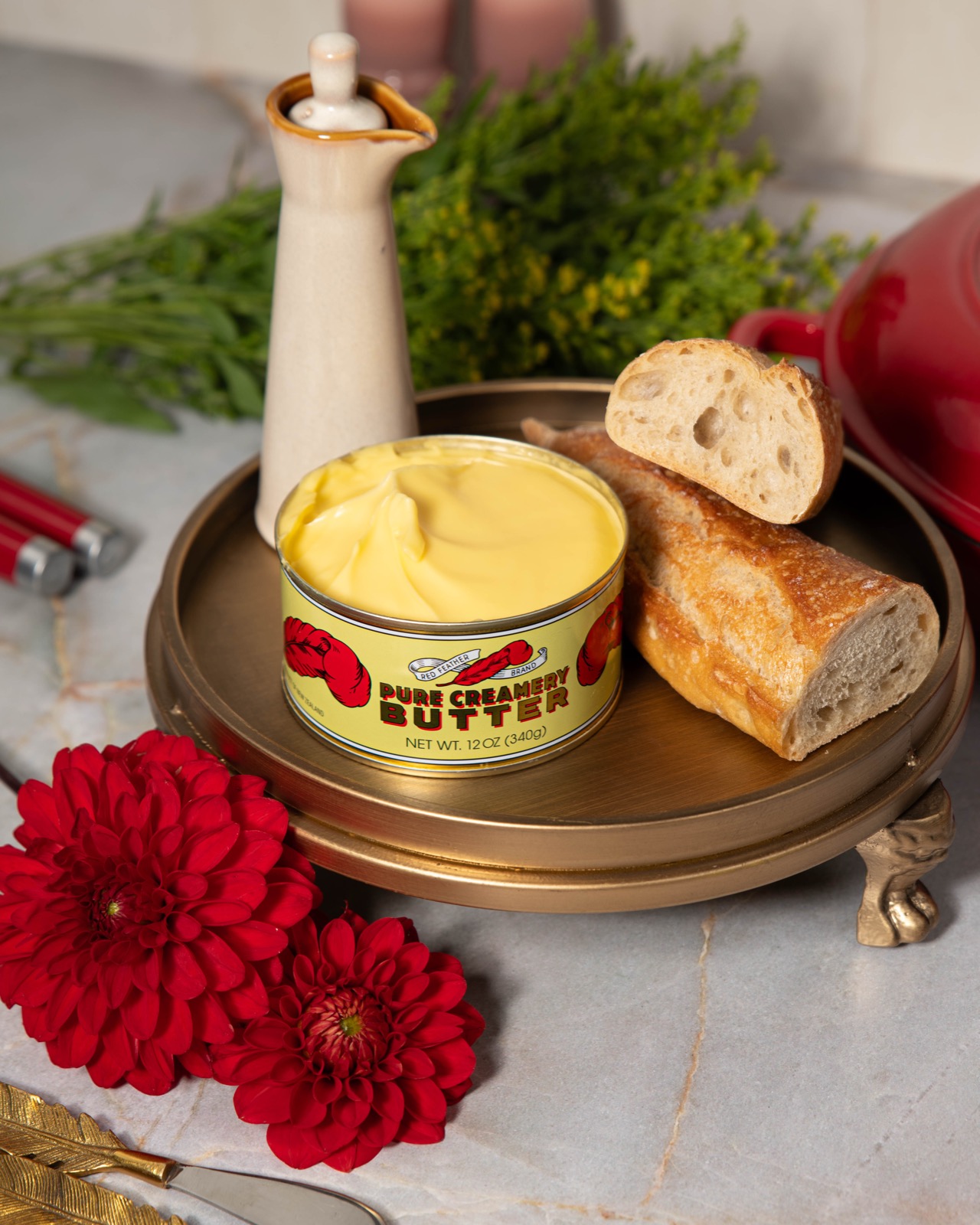 An opened tin of Red Feather butter served on a brass stand with fresh baguette and red dahlias