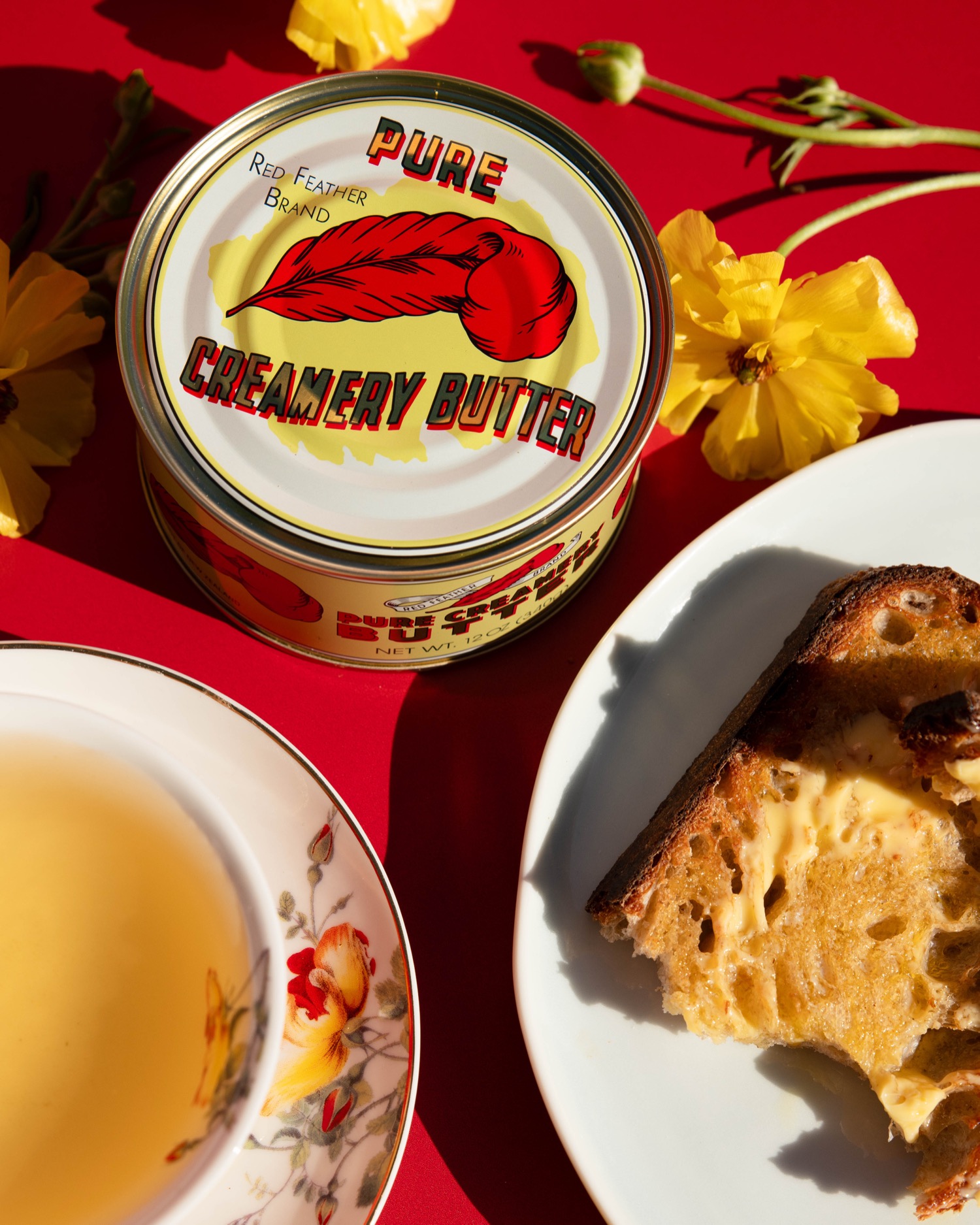 A tin of Red Feather butter beside a cup of tea and buttered toast, seen from above on a red surface