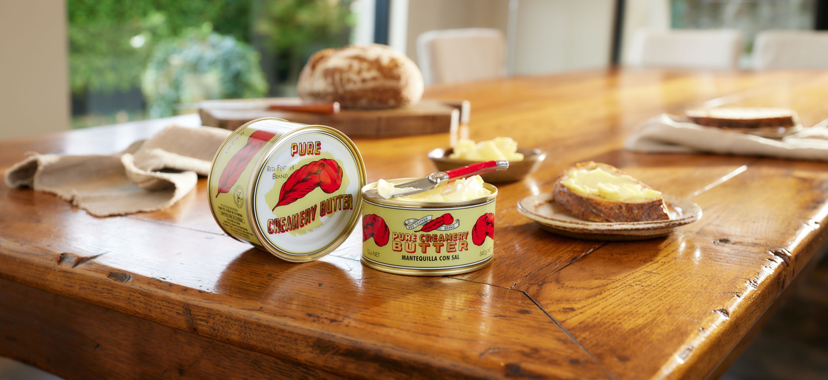 Tins of Red Feather butter on a farmhouse table with sourdough and buttered toast, garden light behind