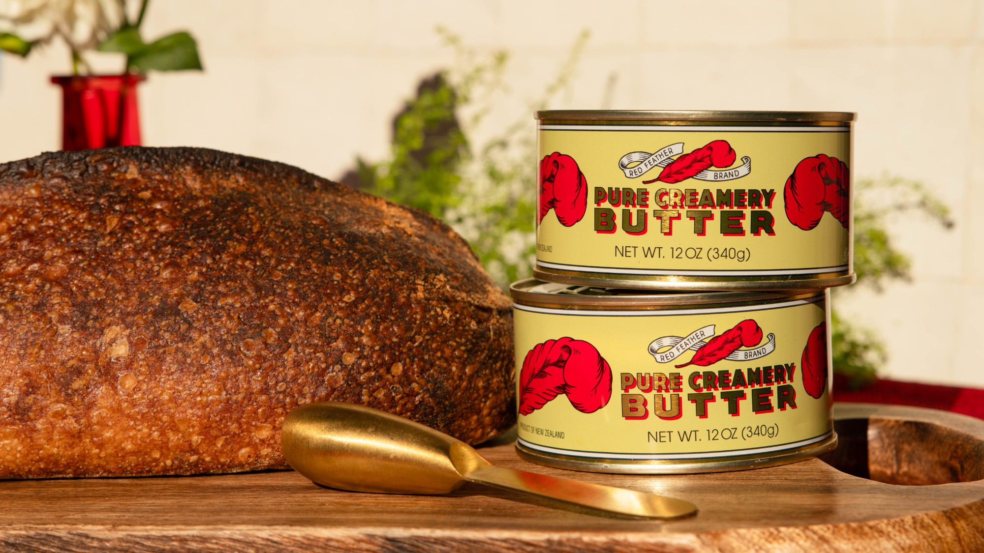 Two tins of Red Feather butter stacked beside a sourdough loaf on a wooden board, garden greenery behind