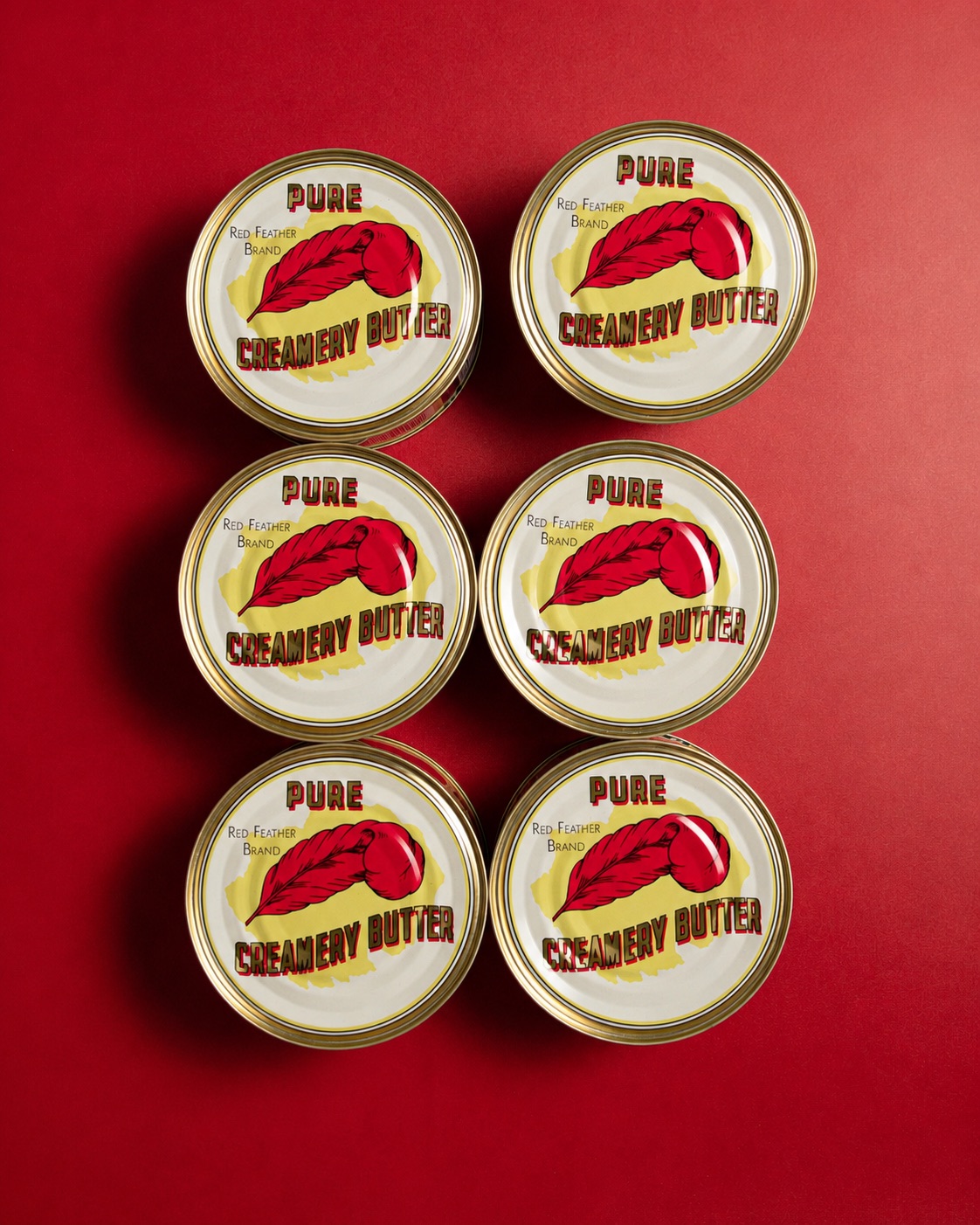 Six 12 ounce tins of Red Feather butter arranged in a grid on a deep red surface, seen from above